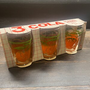 May include: A set of three vintage cola glasses in a box. The clear glasses feature orange slice designs with green leaves. The box is white with a red grid pattern and the text "3 COLA".