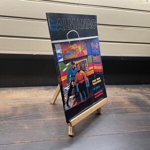 May include: The Franklin Mint Almanac, a vintage book, is displayed on a small wooden easel. The cover features images of Star Trek characters and the text "Star Trek Treasures, TV Toys, Miniature Mansions."