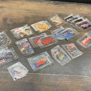 May include: A selection of sealed, vintage stickers with diverse designs, including band logos, celebrity portraits, and graphic art. The stickers are packaged in clear plastic and arranged on a wooden surface.