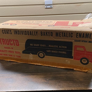 May include: Vintage Structo toy truck box. The cardboard box features red and black graphics of a truck and text that reads "2 Coats Individually-Baked Metallic Enamel." The box is worn, with some tears and creases, suggesting its age.