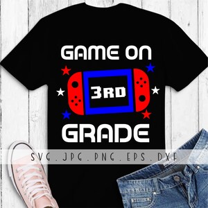 May include: Black t-shirt with the text "GAME ON 3RD GRADE" in white. A red and blue video game controller graphic is in the centre of the shirt. Red and blue stars are around the graphic.