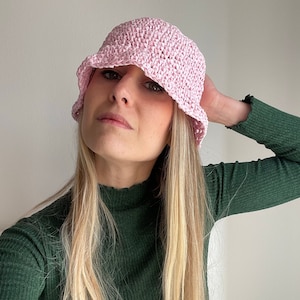 Può includere: Un cappello a secchiello rosa lavorato a uncinetto con un'ampia tesa. Il cappello è indossato da una persona con lunghi capelli biondi e un maglione verde a collo alto.