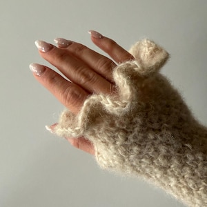 Può includere: Primo piano di un guanto beige senza dita con bordo a balze. Il guanto è realizzato in filato morbido e strutturato. La mano è visibile, con unghie lunghe e curate.