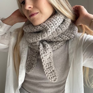 Può includere: Una sciarpa infinita di colore beige chiaro all'uncinetto con un design annodato. La sciarpa è indossata da una persona con lunghi capelli biondi.