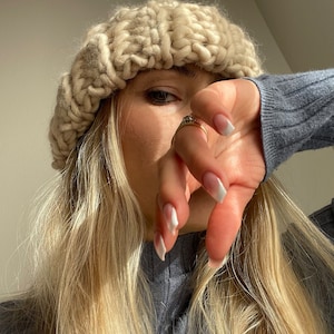 Può includere: Primo piano di una donna che indossa un berretto di lana beige a maglia grossa e un maglione grigio a collo alto. I suoi capelli biondi sono lunghi e fluenti. Indossa un anello al dito e ha le unghie smaltate di bianco.