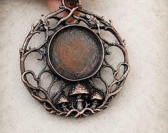 Könnte beinhalten: Ein kreisförmiger Anhänger aus kupferfarbenem Metall mit einem komplizierten durchbrochenen Design aus ineinander verschlungenen Zweigen und Blättern. In der Mitte befindet sich eine runde, vertiefte Fassung. Drei kleine, detaillierte Pilzfiguren befinden sich an der Basis.