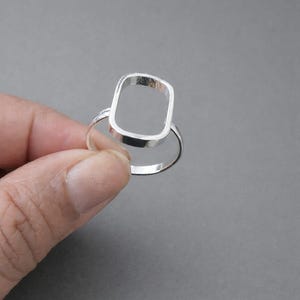 May include: A minimalist silver ring with a rectangular open design. The ring features a smooth, polished finish and a simple band. The geometric shape adds a modern touch to the jewelry piece. The ring is held against a gray background.