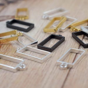 May include: Rectangular metal pendant trays in gold, silver, and black. These open-backed bezels have small loops for attaching to chains or other jewelry components. The trays are scattered on a light brown wooden surface.