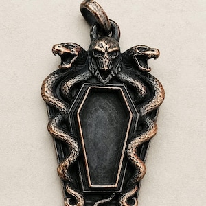 Könnte beinhalten: Ein Metallanhänger mit einer zentralen sargförmigen Vertiefung, die von zwei gewundenen Schlangen und einem Totenkopf an der Oberseite eingerahmt wird. Das Stück hat ein dunkles, oxidiertes Finish mit kupferfarbenen Akzenten auf den Schuppen.