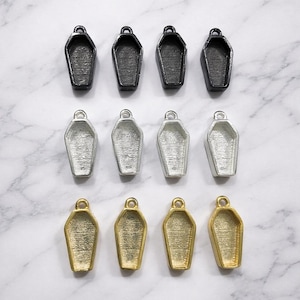 Peut inclure: Vue aérienne de douze breloques en forme de cercueil, disposées en trois rangées. La rangée supérieure est noire, la rangée du milieu argentée et la rangée inférieure dorée. Chaque breloque a une petite boucle en haut pour la fixation.