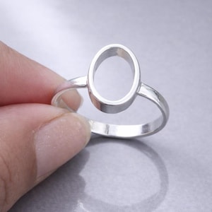 May include: A silver ring featuring an oval-shaped design. The ring is held against a gray background, showcasing its polished surface. The ring's band is thin, and the oval shape is the focal point of the design.