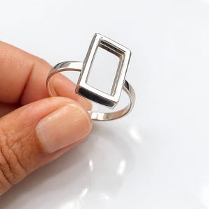 May include: A silver ring featuring a rectangular open frame design. The ring has a polished finish and a simple band. The geometric shape of the frame adds a modern touch to the jewelry.