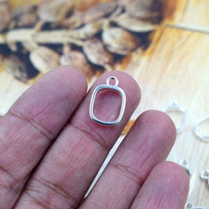 May include: A small, silver-colored rectangular pendant with a loop at the top is held between fingers. Several similar pendants are visible in the background. The pendants are likely for jewelry making.