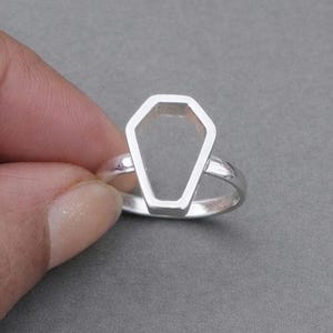 以下が含まれることがあります： 棺の形をしたデザインのシルバーリング。リングは、洗練された仕上がりのミニマリストなオープンフレームスタイルです。リングはグレーの背景に配置され、シンプルでありながらユニークな美しさを表現しています。