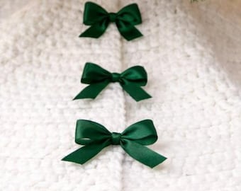 May include: A white, textured Christmas tree skirt with three dark green satin bows. The skirt is beneath a decorated Christmas tree with lights and pine needles. The bows are evenly spaced.