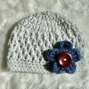 May include: A gray crocheted beanie with a blue flower accent. The flower has a brown button in the center.