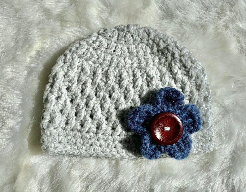 May include: A gray crocheted beanie with a blue flower accent. The flower has a brown button in the center.
