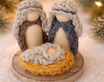 May include: A handcrafted nativity scene featuring two figures with knitted head coverings in brown and blue, and a baby in a yellow and cream-colored knitted cradle, all set on a wooden disc. The background has soft, blurred lights.