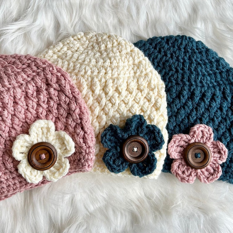 May include: Three crocheted hats with flower embellishments. The hats are in pink, cream, and teal.