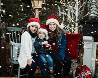 May include: Three children wearing red and white knit Santa hats pose for a Christmas photo. The children are sitting on a wooden chair with a red pillow. The background is a snowy forest with a fireplace and a sign that says "Jingle All The Way".