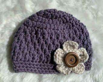 May include: A purple crocheted beanie with a white crocheted flower and a brown button in the center.