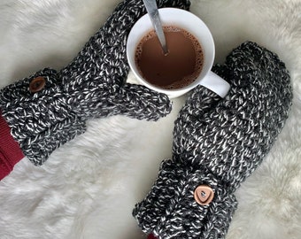 Könnte beinhalten: Ein Paar schwarze und weiße Häkelhandschuhe mit braunen Knöpfen. Die Handschuhe werden von einer Person gehalten, die auch eine weiße Tasse mit heißer Schokolade hält.