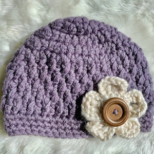 May include: A purple crocheted beanie with a white crocheted flower and a brown button in the center.