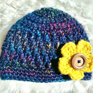May include: A blue crocheted beanie with a yellow crocheted flower and a brown button in the center.