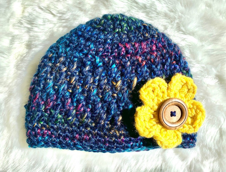 May include: A blue crocheted beanie with a yellow crocheted flower and a brown button in the center.