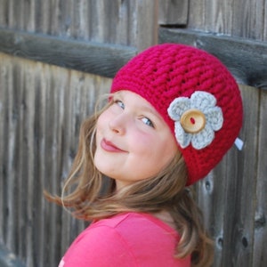 May include: A young girl wearing a pink crocheted hat with a gray flower and a button. The hat has a simple design and is worn slightly off-center.