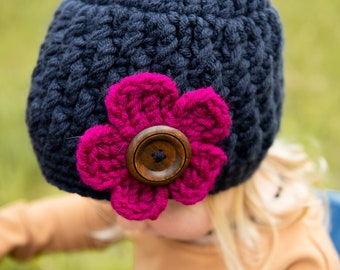 May include: A dark blue crocheted beanie with a pink crocheted flower and a brown button.
