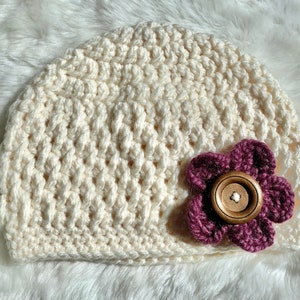 May include: A crocheted cream-colored beanie with a burgundy flower accent. The flower has a brown button in the center.