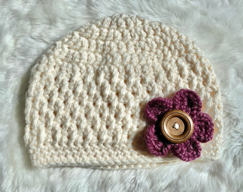 May include: A crocheted cream-colored beanie with a burgundy flower accent. The flower has a brown button in the center.