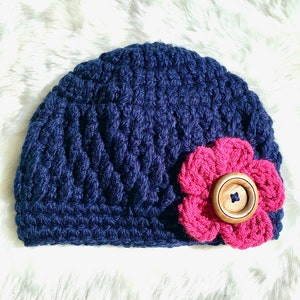 May include: A navy blue crocheted beanie with a pink crocheted flower and a wooden button.