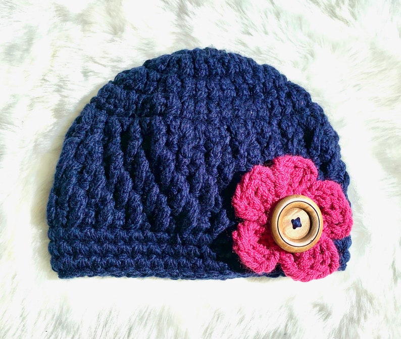 May include: A navy blue crocheted beanie with a pink crocheted flower and a wooden button.