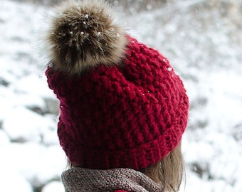 May include: A red crocheted beanie with a brown faux fur pom pom. The beanie is worn by a person in a snowy setting.