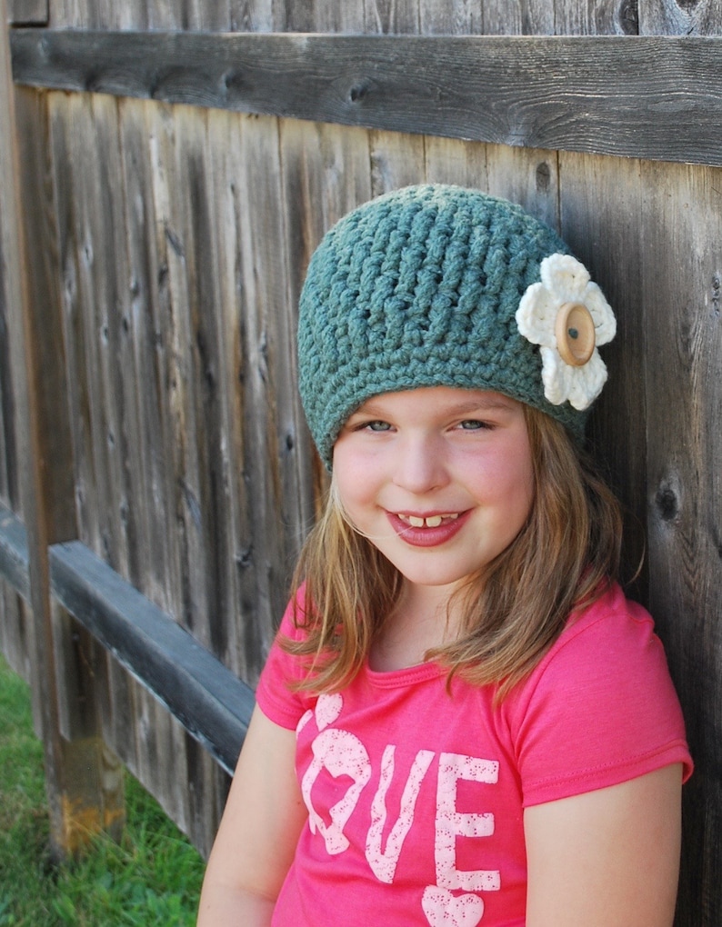 May include: A young girl wearing a teal blue crocheted hat with a white flower and a pink shirt that says "LOVE" on it.