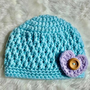 May include: A light blue crocheted baby hat with a purple crocheted heart-shaped embellishment and a wooden button in the center.