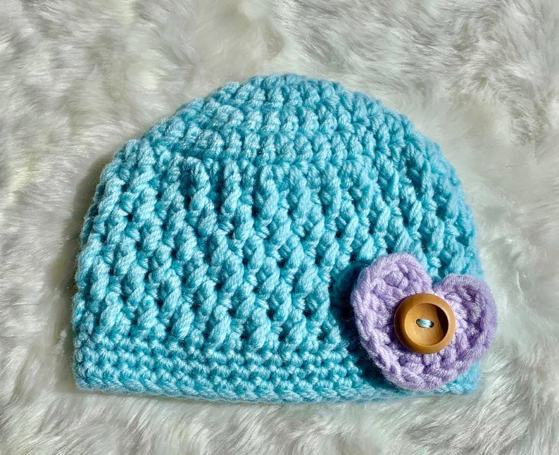 May include: A light blue crocheted baby hat with a purple crocheted heart-shaped embellishment and a wooden button in the center.
