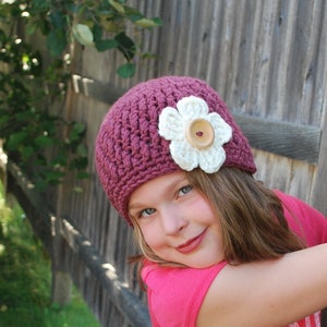 May include: A young girl wearing a burgundy crocheted hat with a white crocheted flower and a brown button in the center. The hat is tilted on her head and she is looking at the camera with a smile.