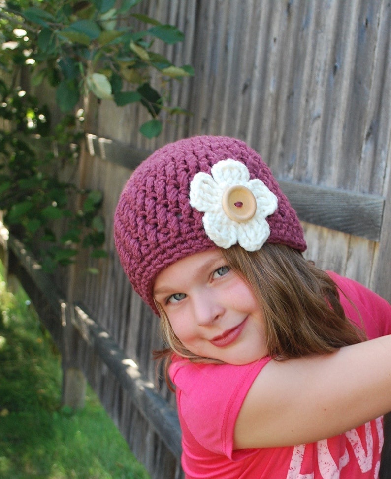 May include: A young girl wearing a burgundy crocheted hat with a white crocheted flower and a brown button in the center. The hat is tilted on her head and she is looking at the camera with a smile.