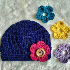 May include: A blue crocheted beanie with a pink crocheted flower and a wooden button. The beanie is surrounded by three crocheted flowers in teal, yellow, and purple.
