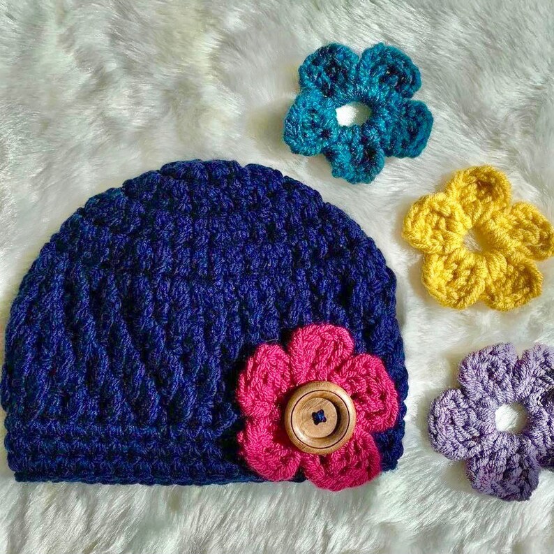 May include: A blue crocheted beanie with a pink crocheted flower and a wooden button. The beanie is surrounded by three crocheted flowers in teal, yellow, and purple.
