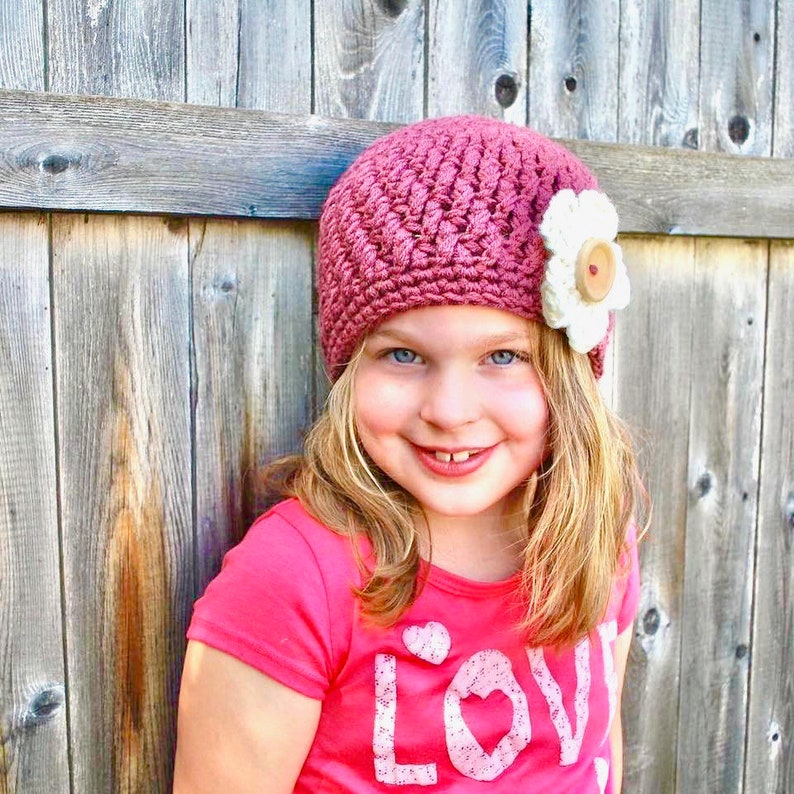 May include: A young girl wearing a pink crocheted hat with a white flower and a wooden button. She is wearing a pink shirt with the word "LOVE" on it.