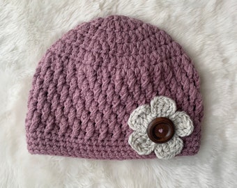 May include: A mauve crocheted beanie with a white crocheted flower and a brown button in the center.