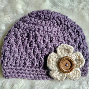 May include: A purple crocheted beanie with a white crocheted flower and a brown button in the center.