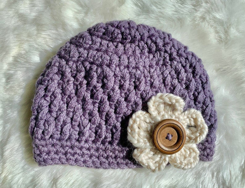 May include: A purple crocheted beanie with a white crocheted flower and a brown button in the center.