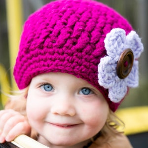 May include: A young child wearing a pink crocheted hat with a purple flower and a brown button. The hat has a textured design.