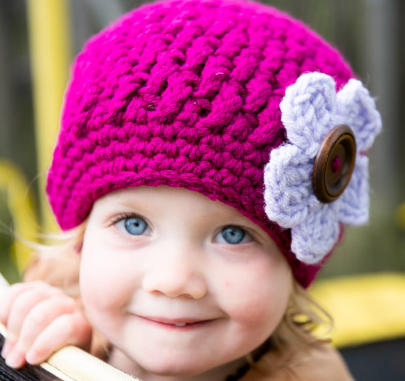 May include: A young child wearing a pink crocheted hat with a purple flower and a brown button. The hat has a textured design.