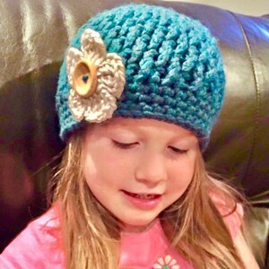 May include: A young girl wearing a teal blue crocheted hat with a white crocheted flower and a brown button. The hat is worn on her head and she is looking down.